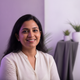 Portrait of a smiling South Asian woman in her late 30s, with a soft lavender and white background.